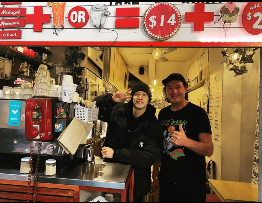 Bang Chan of Stray Kids posing with another person behind the Jungle Juice Bar counter